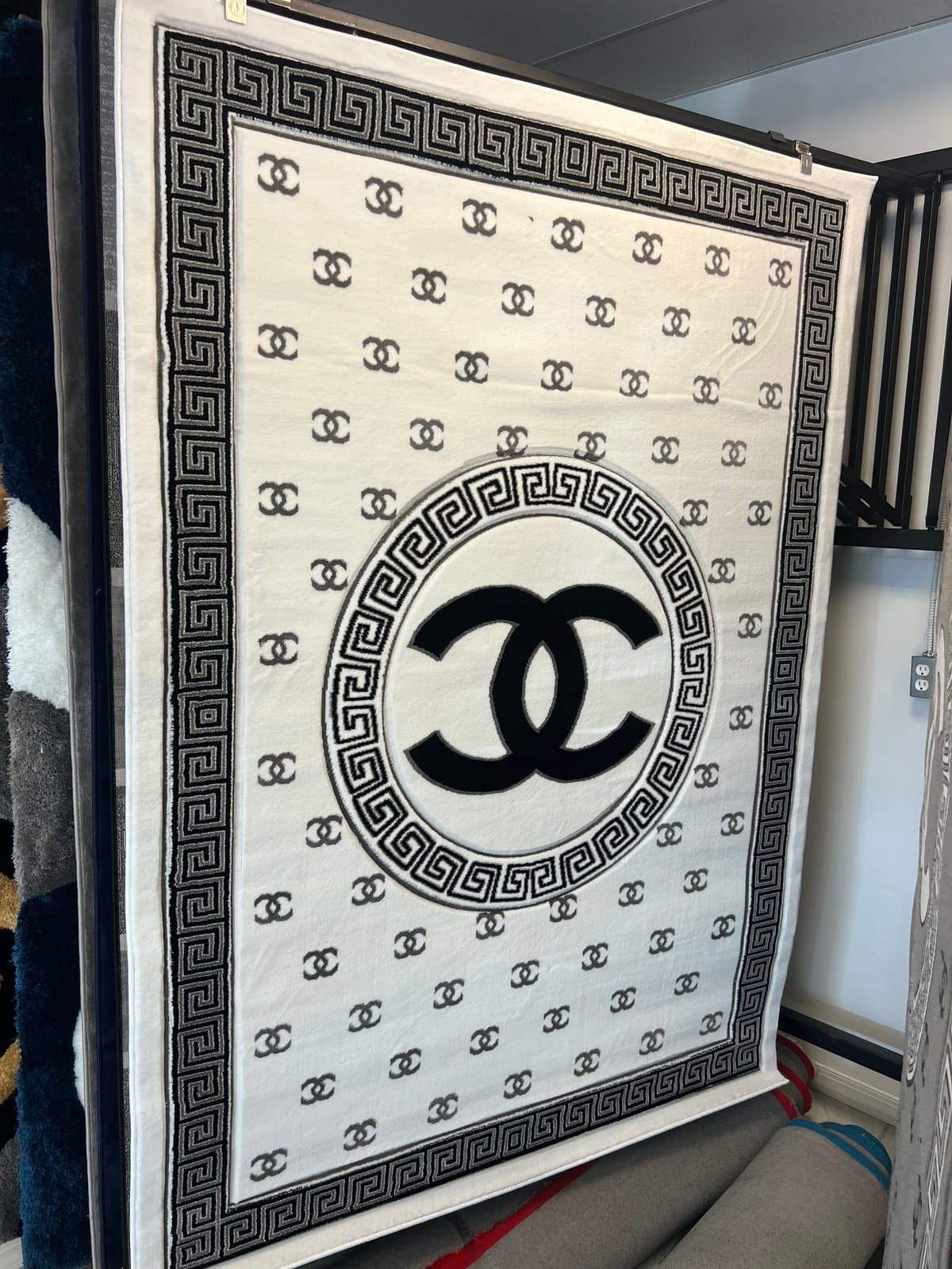 Luxury designer blanket with cream background, repeating interlocking CC logo pattern, black geometric Greek key border, and large centered black CC double-C logo